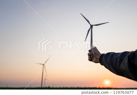 Man holds toy wind turbine in his hand. Real wind turbines in the background Man holds toy wind turbine in his hand. Real wind turbines in the background 119763877