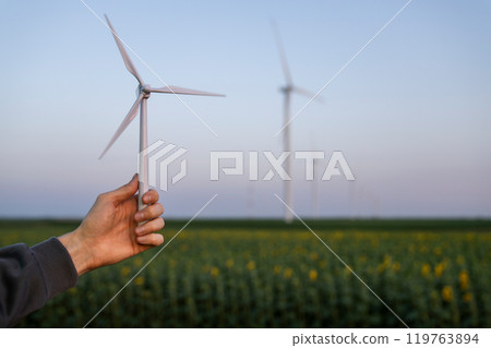 Man holds toy wind turbine in his hand. Real wind turbines in the background 119763894