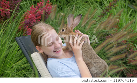 A joyful young woman holds her rabbit close while relaxing on a lounge chair, sharing a happy moment outdoors. A joyful young woman holds her rabbit close while relaxing on a lounge chair, sharing a happy moment outdoors. 119764119