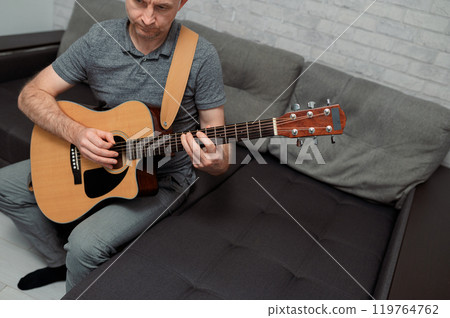 Guitarist paying a acoustic guitar in office. portrait Male musician sitting on sofa and playing music instrument Guitarist paying a acoustic guitar in office. portrait Male musician sitting on sofa and playing music instrument 119764762