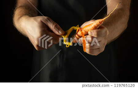Vegetable peeler in the hand of a cook to clean carrots before preparing a vitamin salad. Low key concept of preparing vegetables for dinner 119765120