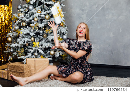 Blonde woman in dress smiling with gifts at Christmas tree for new year 119766284