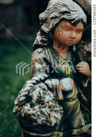 Worn stone angel. Vintage female figurine with shabby paint on her face. Monument in a cemetery. Old statue close up. Sadness, longing, depression. Worn stone angel. Vintage female figurine with shabby paint on her face. Monument in a cemetery. Old statue close up. Sadness, longing, depression. 119766384