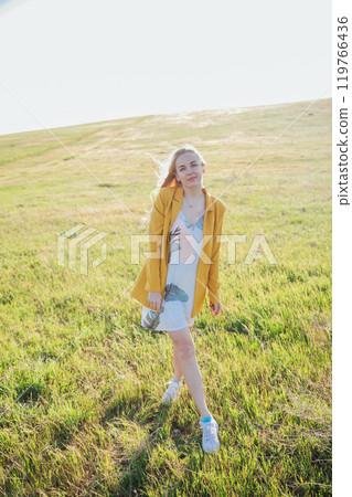 Beautiful blonde woman in summer dress walking in blooming field at sunset Beautiful blonde woman in summer dress walking in blooming field at sunset 119766436