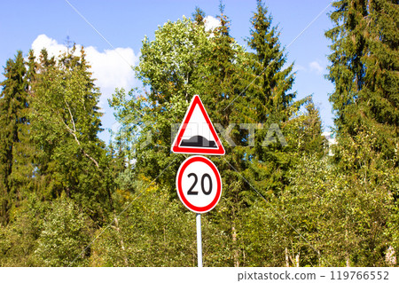 Two road signs for motorists warning about pedestrians and cyclists - speed limit to 20 kmh per hour, a sign of a speed bump on the background of green pine forests and blue sky. Caution speed bumps. Two road signs for motorists warning about pedestrians and cyclists - speed limit to 20 kmh per hour, a sign of a speed bump on the background of green pine forests and blue sky. Caution speed bumps. 119766552