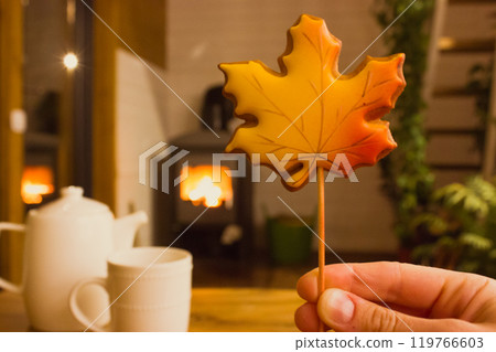 Gingerbread cookie in shape of autumnal orange maple leaf in woman's hand against burning fire in fireplace in cozy comfort home in the evening. Tea cup, teapot on a wooden table in dark room. 119766603