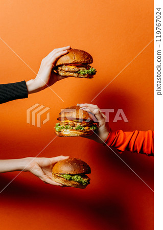 Three hands holding delicious burgers on vibrant background, food advertisements and promotions 119767024