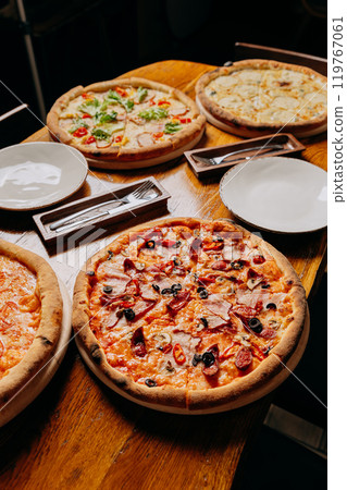 Assorted pizzas on a rustic wooden table, ready to serve with plates and cutlery Assorted pizzas on a rustic wooden table, ready to serve with plates and cutlery 119767061