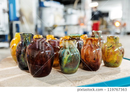 Many colorful glass vases at glass blowing manufacture factory  with furnace equipment set on background. Handmade handcrafted blowing glassware design production workshop Europe 119767363