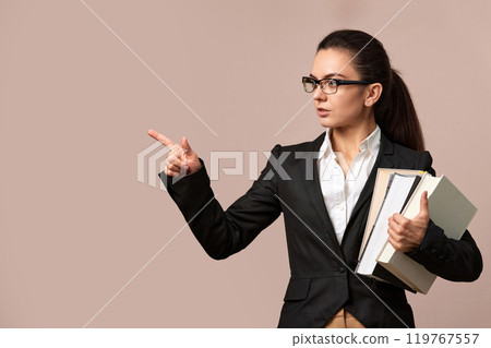 attractive woman in glasses and black jacket holding stack of books and pointing at the side, education concept. 119767557