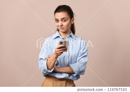 skeptical pretty woman holding mobile phone looking at camera on beige background. 119767583