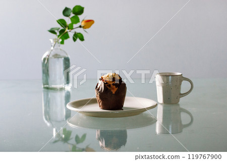 A cupcake on a white plate and a glass vase with a yellow rose, a small beige cup for tea or coffee. Still life with food and drink. Chocolate maffin or muffin - traditional American dessert. A cupcake on a white plate and a glass vase with a yellow rose, a small beige cup for tea or coffee. Still life with food and drink. Chocolate maffin or muffin - traditional American dessert. 119767900