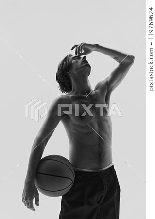 Young man holding basketball, hand raised to his face in thoughtful pose. Dynamic photo in monochrome filter. Young man holding basketball, hand raised to his face in thoughtful pose. Dynamic photo in monochrome filter. 119769624