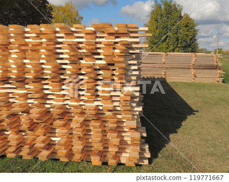 Wooden narrow planks stacked on top of each other lie on grass against background of deciduous trees and cloudy sky, building materials from fresh wood outdoor storage 119771667
