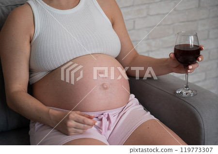 Close-up of the belly of a pregnant woman holding a glass of red wine while sitting on the sofa. Skin rash.  119773307