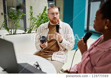 Medium shot of male podcast guest talking to female host while recording new episode holding professional microphone sitting at cafe table, copy space Medium shot of male podcast guest talking to female host while recording new episode holding professional microphone sitting at cafe table, copy space 119773569