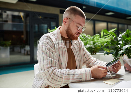Medium shot of handsome man wearing glasses to read tablet screen while planning work or studying remotely at table in cozy sunlit cafe in modern business center, copy space 119773624