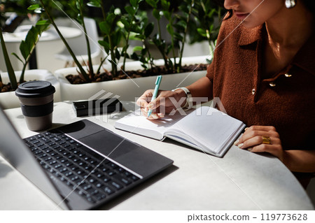 Cropped shot of unrecognizable businesswoman writing in notebook while working on new project or studying at table in cozy sunlit cafe, copy space Cropped shot of unrecognizable businesswoman writing in notebook while working on new project or studying at table in cozy sunlit cafe, copy space 119773628