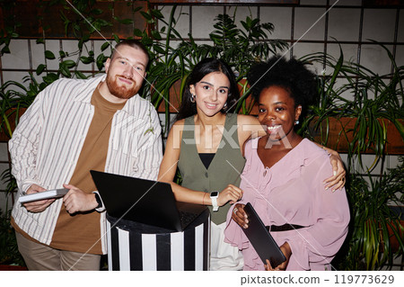 Portrait of three smiling business people from different ethnic backgrounds collaborating in cafe with green living wall, working on laptop and digital tablet in modern coworking space 119773629