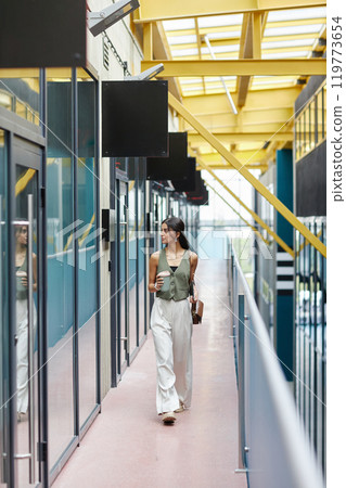 Vertical shot of fashionable businesswoman dressed in elegant waistcoat and white trousers walking with reusable cup of coffee towards office room in business building, copy space Vertical shot of fashionable businesswoman dressed in elegant waistcoat and white trousers walking with reusable cup of coffee towards office room in business building, copy space 119773654