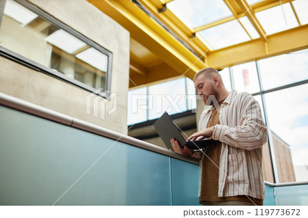 Side view of adult man wearing casual clothes focused on using laptop while standing on business center balcony Side view of adult man wearing casual clothes focused on using laptop while standing on business center balcony 119773672