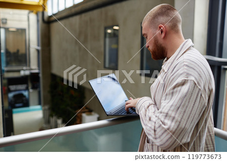 Side view of adult businessman in casual outfit concentrated on using laptop standing on balcony of business center, copy space Side view of adult businessman in casual outfit concentrated on using laptop standing on balcony of business center, copy space 119773673