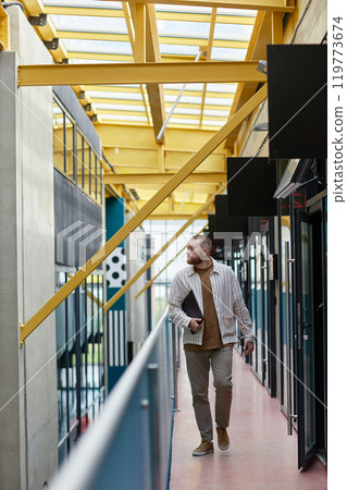 Vertical shot of successful red bearded office worker dressed in casual clothes holding laptop walking past premises of two storey business center, copy space Vertical shot of successful red bearded office worker dressed in casual clothes holding laptop walking past premises of two storey business center, copy space 119773674