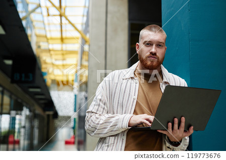 Medium shot of adult handsome man holding open laptop looking at camera while standing next to blue column in office center, copy space 119773676