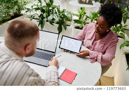Smiling African American businesswoman holding digital tablet with white mockup screen presenting male colleague work project at business meeting in cafe at modern office center 119773683