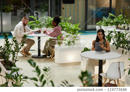 Wide angle shot of woman using tablet while sitting at table in cafe of office building. Business people working in cozy cafe featuring modern biophilic design Wide angle shot of woman using tablet while sitting at table in cafe of office building. Business people working in cozy cafe featuring modern biophilic design 119773686