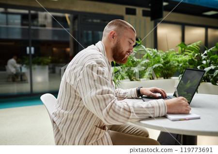 Side view of concentrated adult man using laptop and writing in notebook while studying or working at table in office center cafe, copy space 119773691