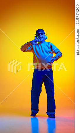 Full-length portrait of stylish elderly bearded man, in sunglasses and cap, listening to music in headphones and dancing against yellow studio background in neon light 119774209