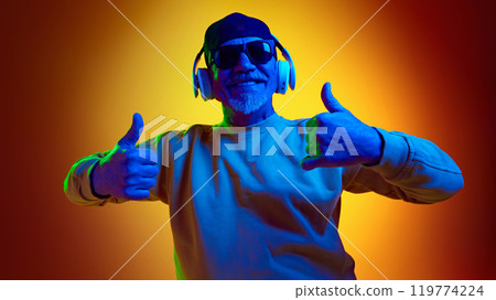 Positive elderly man in sunglasses listening to music in headphones against yellow studio background in neon light Positive elderly man in sunglasses listening to music in headphones against yellow studio background in neon light 119774224