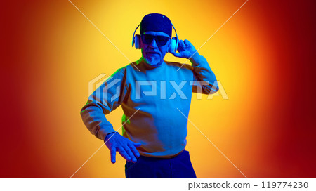Stylish dj. Elderly bearded man in sunglasses, dj, listening to music in headphones against yellow studio background in neon light 119774230