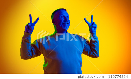 Portrait of happy elderly man posing with funny happy face, showing peace gesture and smiling against yellow studio background in neon light Portrait of happy elderly man posing with funny happy face, showing peace gesture and smiling against yellow studio background in neon light 119774234