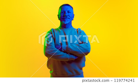 Portrait of stylish, bearded, serious elderly man confidently posing, looking aside against yellow studio background in neon light 119774241