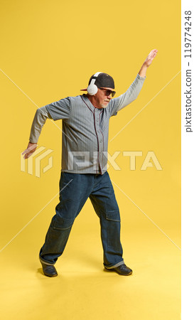 Full-length image of elderly bearded man in cap and sunglasses listening to music in headphones and dancing against yellow studio background 119774248