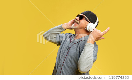 Elderly bearded man in cap and sunglasses listening to music in headphones against yellow studio background. Enjoyment 119774263