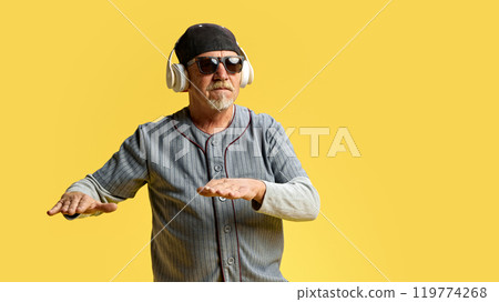 Elderly bearded man in cap and sunglasses listening to music in headphones against yellow studio background. Stylish dj Elderly bearded man in cap and sunglasses listening to music in headphones against yellow studio background. Stylish dj 119774268
