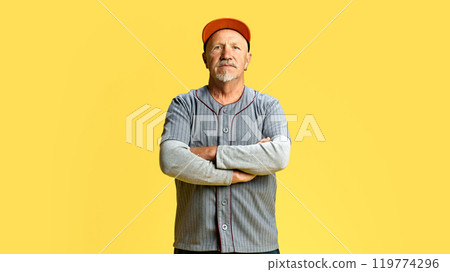 Portrait of senior man standing with serious but calm expression, wearing grey shirt and cap, against yellow studio background Portrait of senior man standing with serious but calm expression, wearing grey shirt and cap, against yellow studio background 119774296