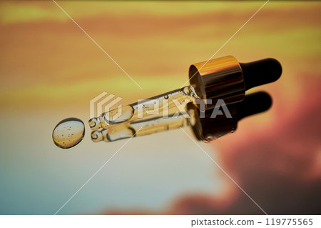 A pipette with facial serum against a sunset background in bright orange tones. 119775565