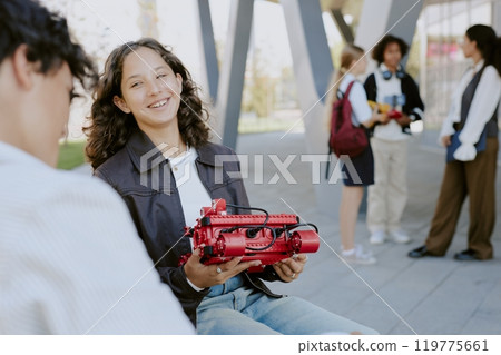 Caucasian curly haired teen girl holding red robot and looking at her male classmate while they having robotics open air lesson Caucasian curly haired teen girl holding red robot and looking at her male classmate while they having robotics open air lesson 119775661