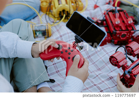 High angle shot of unrecognizable boy sitting on blanket surrounded with multicolored robots holding plastic red joystick with set smartphone 119775697