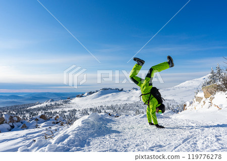 Man stands on hands Bright acid green outfit: warm suit, goggles, black helmet. snowy mountains background ski resort. 119776278