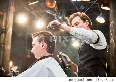 Professional barber using hairspray to finish hairstyle. Stylish hairdresser making final touches. Young customer getting fresh haircut. Professional barber using hairspray to finish hairstyle. Stylish hairdresser making final touches. Young customer getting fresh haircut. 119776294