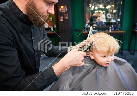 Cute boy excited about new haircut. Blond boy getting stylish haircut in barbershop. Professional hairdresser holding shaver and comb. 119776296