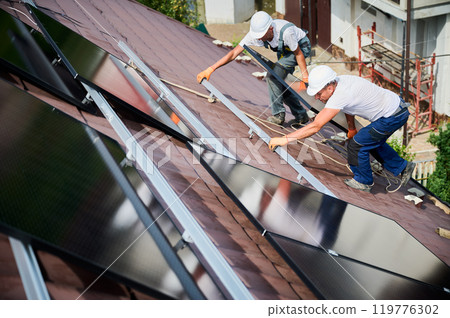 Workers building solar panel system on roof of house. Two men installers in helmets carrying photovoltaic solar module outdoors. Alternative, green and renewable energy generation concept. Workers building solar panel system on roof of house. Two men installers in helmets carrying photovoltaic solar module outdoors. Alternative, green and renewable energy generation concept. 119776302