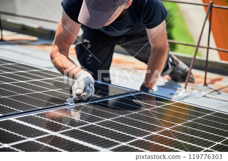 Worker building photovoltaic solar panel system on rooftop of house. Close up of man engineer in gloves installing solar module with help of wrench outdoors. Alternative energy generation concept. 119776303