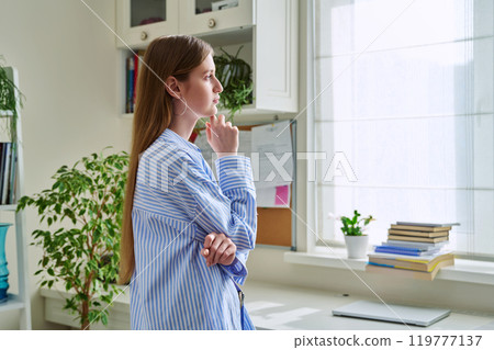 Serious young woman looking out the window, in home interior 119777137
