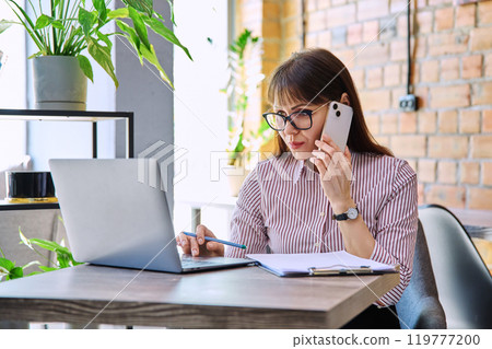 Portrait off middle-aged business woman working with laptop, talking on phone, sitting at desk Portrait off middle-aged business woman working with laptop, talking on phone, sitting at desk 119777200